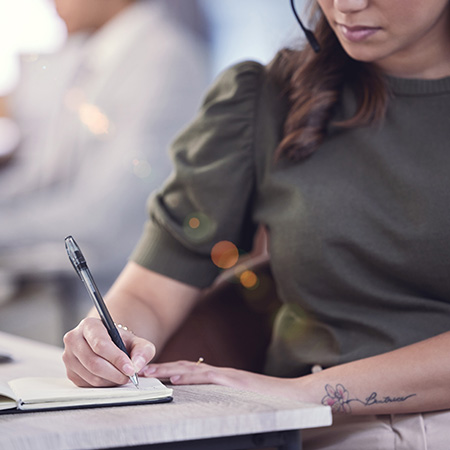 woman writing in notebook at desk in a professional setting with headset and floral tattoo focusing on organization and productivity techniques for success with 13 strategies